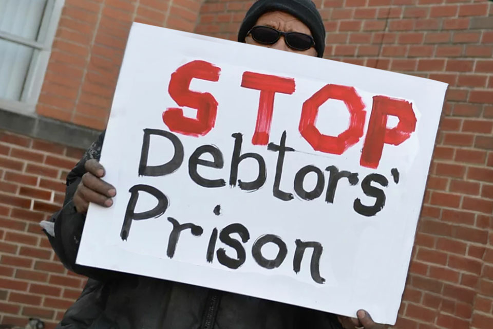 A male demonstrator standing outside a brick building holds up a sign saying, “Stop Debtors’ Prison.” 